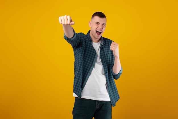 Emotional young man shouting celebrating victory shaking fists yellow background