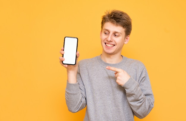 Emotional young man standing on yellow holds a smartphone in his hands and looks at the white screen, showing a finger