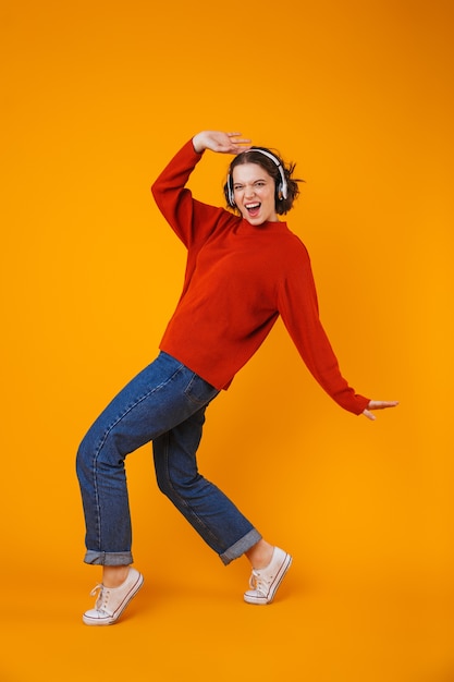 Emotional young pretty woman posing isolated on yellow wall listening music with headphones dancing.