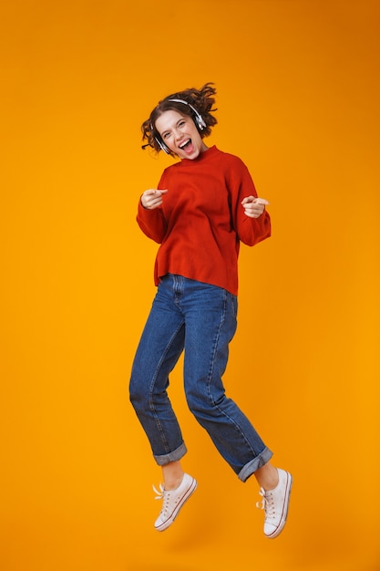 emotional young pretty woman posing isolated on yellow wall listening music with headphones jumping.