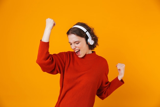 emotional young pretty woman posing isolated on yellow wall listening music with headphones make winner gesture.