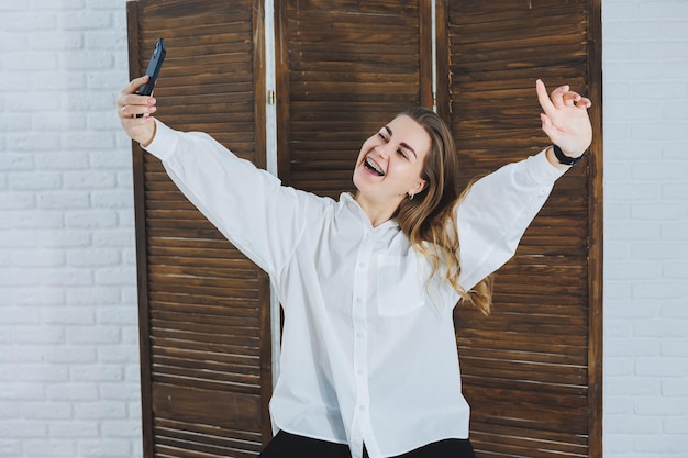 Emotional young woman in a white cotton shirt gesticulates and looks at the screen of a modern gadget A girl with a mobile phone takes a selfie