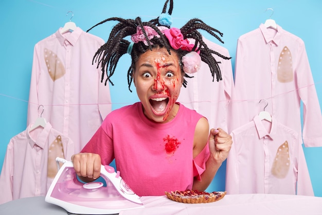 Emotional young woman with braided hairstyle face smeared with\
sweet jam eats delicious pie irons laundry at home screams loudly\
clenches fist has much work to do housekeeping routines\
concept