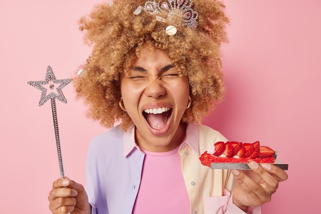 Фото Эмоциональная молодая женщина с вьющимися густыми волосами держит небольшой вкусный клубничный торт и клубничный торт, одетый в повседневную рубашку, изолированную на розовом фоне концепция праздника людей
