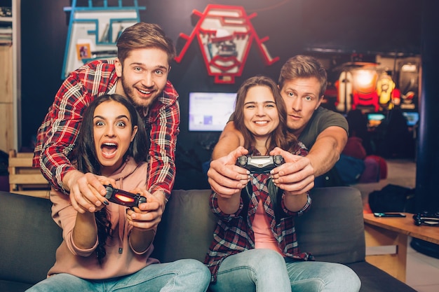 Emotional young women sit on sofa in playing room. They hold gamepads and playing. Guys help them.