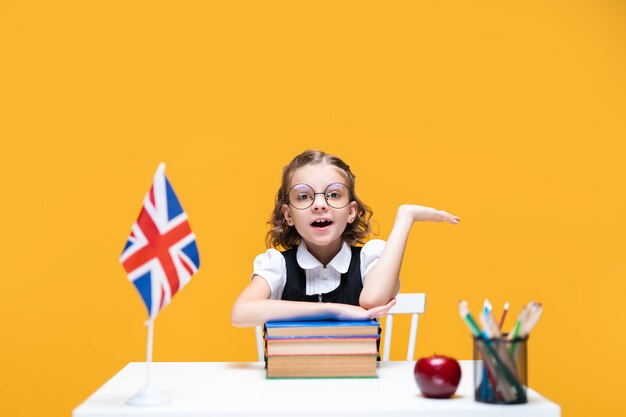 Emotioneel kaukasisch schoolmeisje zit aan de balie en steekt hand engelse les op met de vlag van groot-brittannië