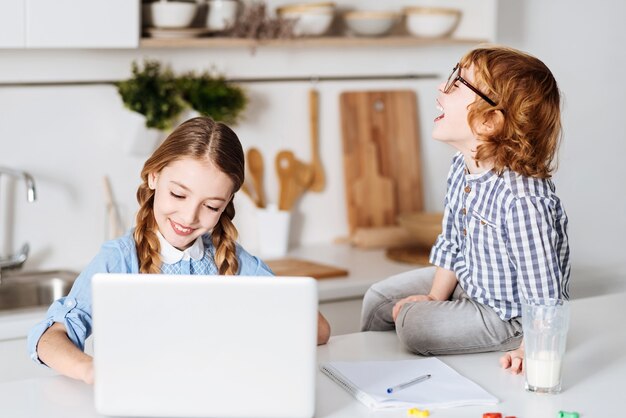 Emotioneel kind. Nieuwsgierige, humoristische, slimme jongen die behoorlijk opgetogen kijkt over wat zijn zus zegt tijdens het studeren met haar laptop