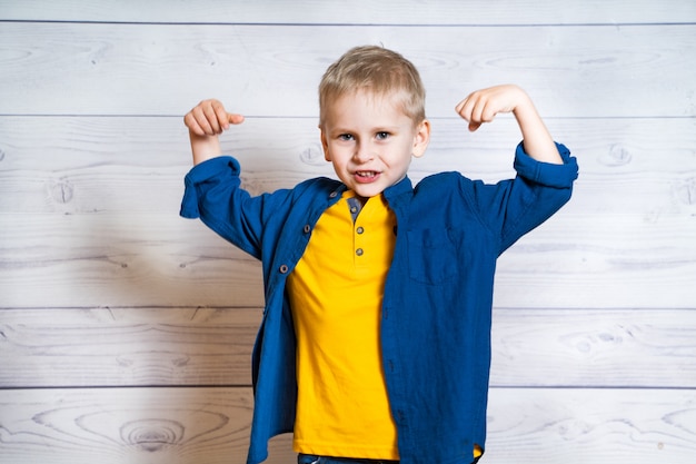 Emotioneel portret van een kleine blonde jongen die blauw overhemd draagt. Leuk jong geitje dat sterkte toont door zijn handen terwijl het kijken