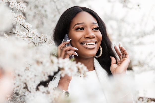 Emotioneel portret van een mooie jonge zwarte vrouw met een glimlach in de buurt van bloemen praten op een slimme telefoon buitenshuis. Gelukkig lachend Afrikaans meisje geniet van een mobiele communicatie aan de telefoon in de buurt van lentebomen