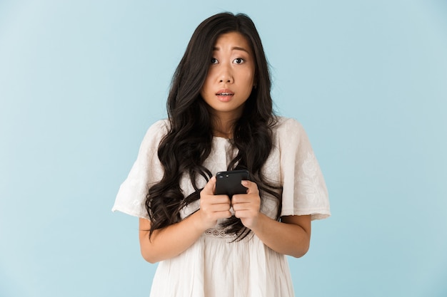 Emotionele Aziatische mooie vrouw geïsoleerd over blauwe muur met behulp van mobiele telefoon