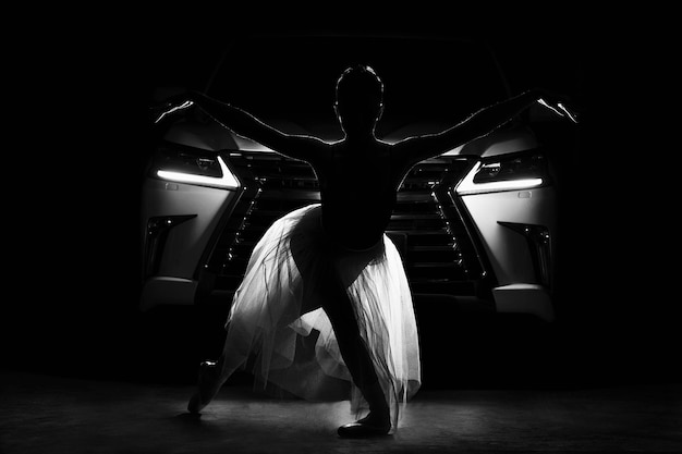 Emotionele ballerina in een doorschijnende rok poseert tegen het silhouet van een auto