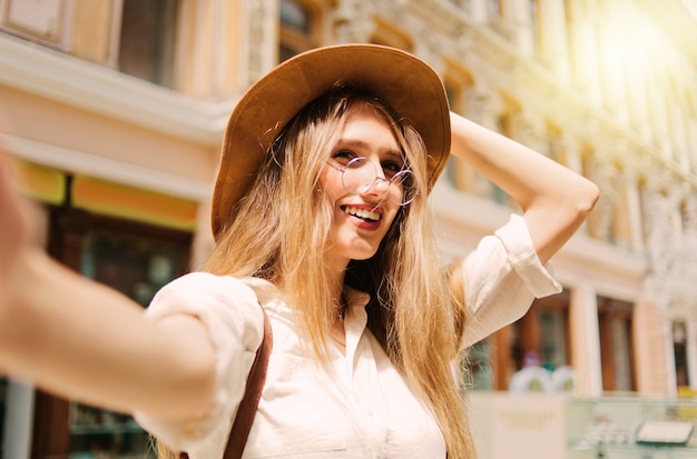 Emotionele blonde vrouw in vilten hoed en bril maakt selfie oude stadsarchitectuur