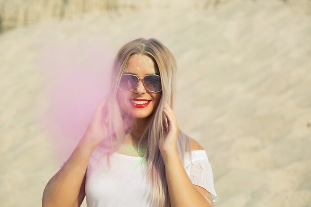 Emotionele blonde vrouw met een shirt met blote schouders en een bril die speelt met roze droge verf Holi in de woestijn