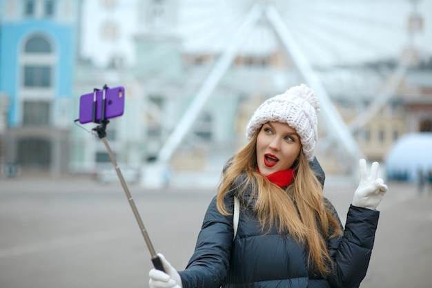 Emotionele blonde vrouw toerist met gebreide muts, selfie nemen op straat in de winter. Ruimte voor tekst