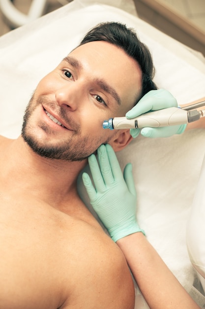 Foto emotionele donkerharige man die lacht terwijl schoonheidsspecialist in rubberen handschoenen zijn gezicht aanraakt tijdens de voedende procedure