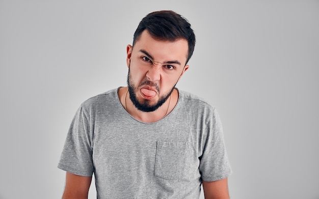 Emotionele en bebaarde jonge knappe brunette man grimassen, mond maken, zijn tong naar de camera uitsteken om iemand te plagen, zich gedragen als een klein ondeugend kind.