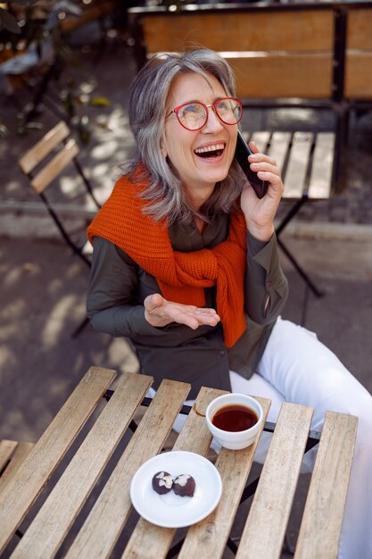 Emotionele grijsharige vrouw praat aan de telefoon aan tafel op het terras van een café