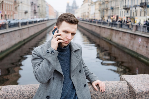 Emotionele jonge man met mobiele telefoon die een elegante grijze jas draagt