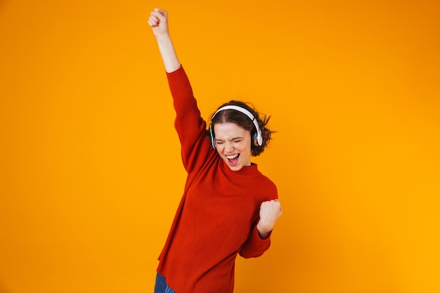 emotionele jonge mooie vrouw poseren geïsoleerd op gele muur luisteren muziek met koptelefoon maken winnaargebaar.