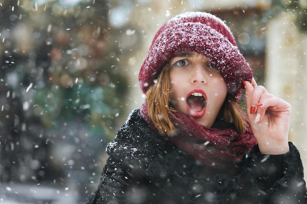 Emotionele jonge vrouw draagt een rode dop die tijdens sneeuwval door de stad loopt. Lege ruimte