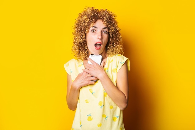 Emotionele mooie krullende jonge vrouw met een telefoon op een gele achtergrond