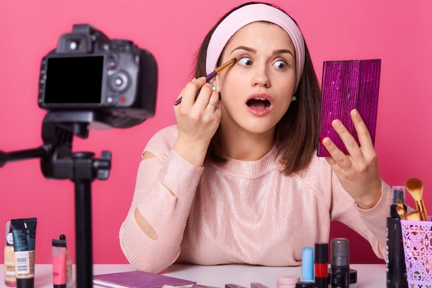 Emotionele mooie make-up artiest met wijd geopende mond met roze spiegel in één hand, oogschaduw aanbrengen met professionele borstel, video opnemen voor haar nieuwe vlog.