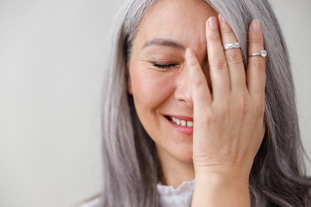 Emotionele portretten van een verbleekte langharige Aziatische gerijpte vrouw op grijze muur