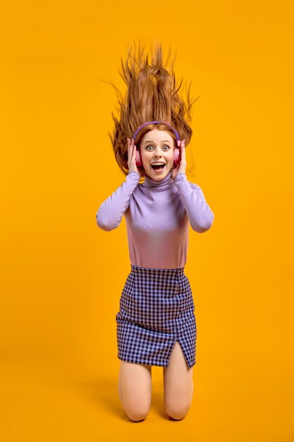 Emotionele vrolijke gekke vrouw in koptelefoon met vliegende haren geïsoleerd op gele kleur achtergrond in studio, portret. Opgewonden blanke vrouw met natuurlijk rood haar in shirt en rok geniet van muziek