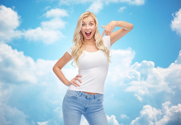 эмоции, выражения, реклама и концепция людей - счастливая улыбающаяся молодая женщина или девочка-подросток в белой футболке, указывающая пальцем на себя на фоне голубого неба и облаков