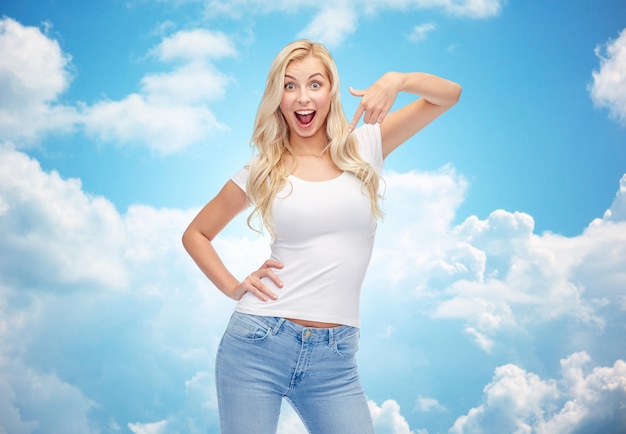 emotions, expressions, advertisement and people concept - happy smiling young woman or teenage girl in white t-shirt pointing finger to herself over blue sky and clouds background