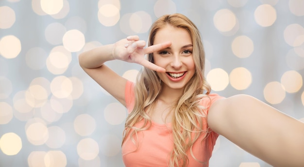 emotions, expressions and people concept - happy smiling young woman taking selfie and showing peace hand sign over holidays lights background