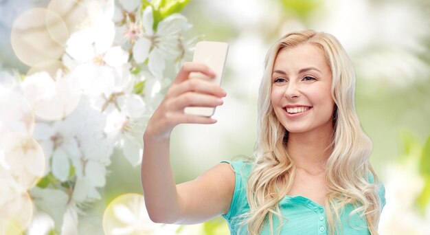 Photo emotions, expressions and people concept - happy smiling young woman or teenage girl taking selfie with smartphone over natural spring cherry blossom background