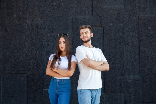 Emotions gesture expression and people concept Serious Caucasian man and woman in white Tshirts and jeans standing next to each other and crossing their arms on a black background with copy space