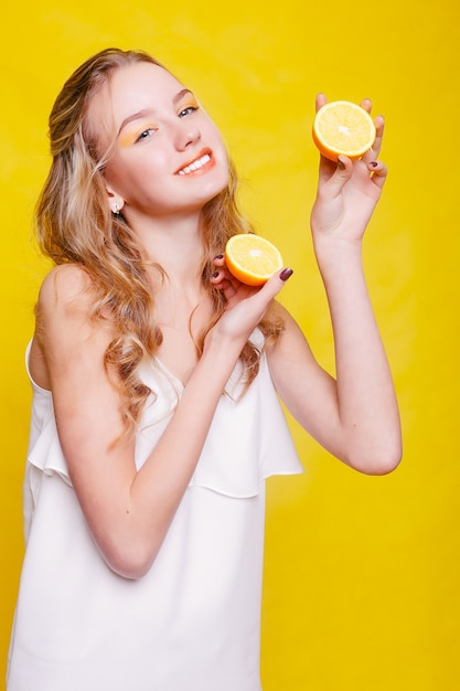 Emotions, health, people, food and beauty concept - Beauty Model Girl takes Juicy Oranges. Beautiful Joyful teen girl with freckles, funny hairstyle. yellow makeup. Professional make up. Orange Slice