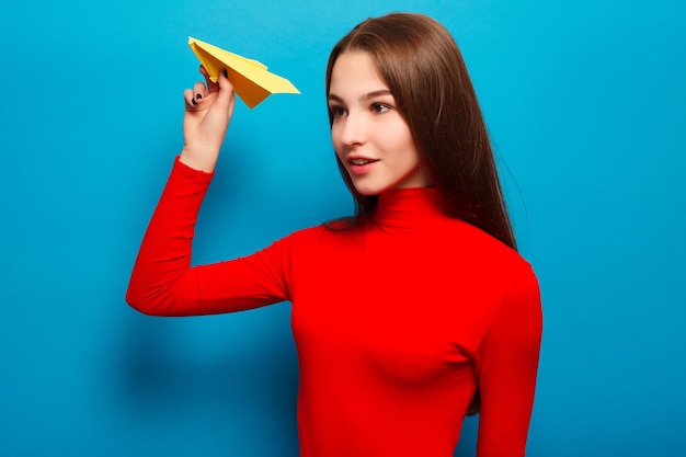 Emotions, people, beauty, fashion and lifestyle, travel, tourism and people concept - Portrait of a beautiful woman holding a yellow paper airplane. On a blue background.