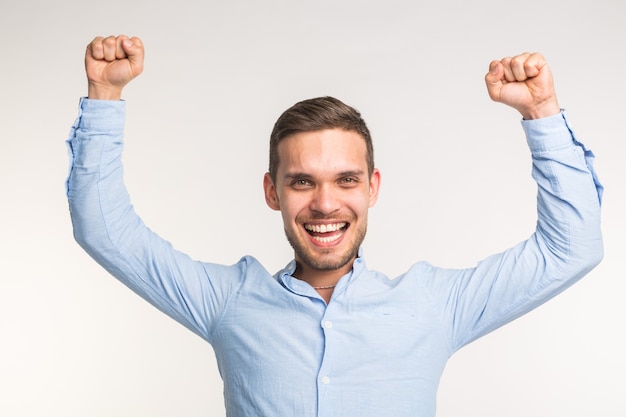 Emotions and people concept - young happy man raised his fists up over the white wall.