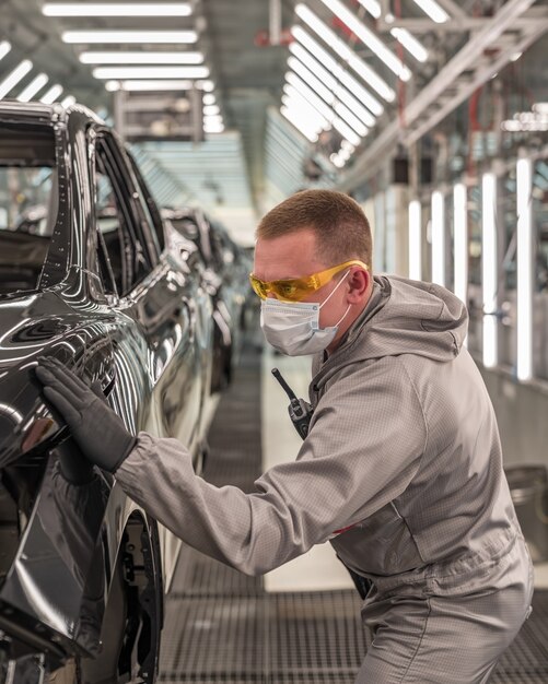 An employee of an automobile factory wearing a mask checks the quality of the painted surface