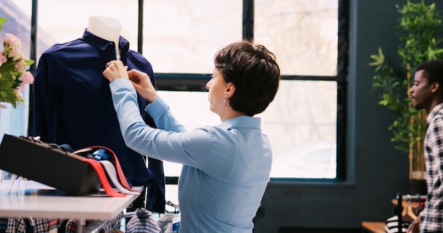 Employee closing shirt buttons, arranging stylish merchandise in modern boutique. Caucasian manager working at store visual, checking hangers full with new fashion collection in shopping mall