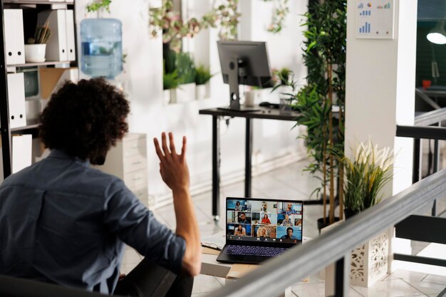 Impiegato in una videoconferenza di gruppo, che saluta diversi colleghi sullo schermo del portatile. manager esecutivo e team remoto che collaborano al progetto durante una riunione online virtuale nello spazio di lavoro aziendale