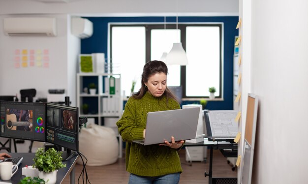 Employee holding laptop standing in creative agency office prosessing customer video project in post production software