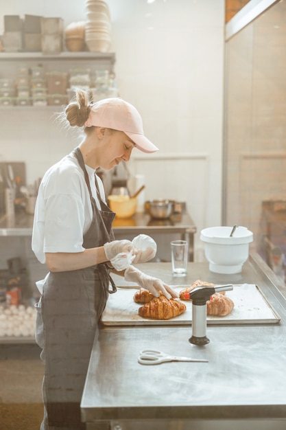 Employee holds confectioner sleeve with delicious cream to decorate croissants in bakery shop