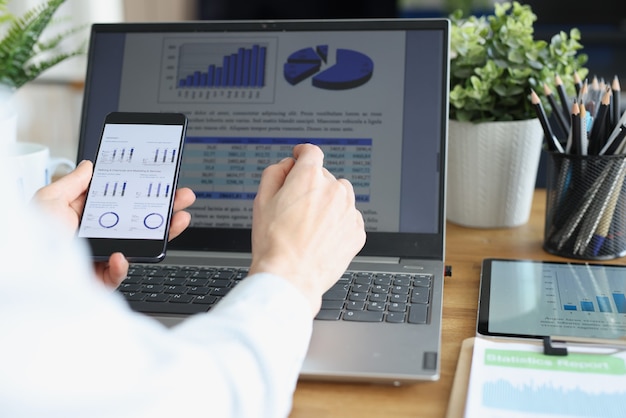 Employee holds smartphone with commercial business charts in his hand and compares them with image on laptop monitor