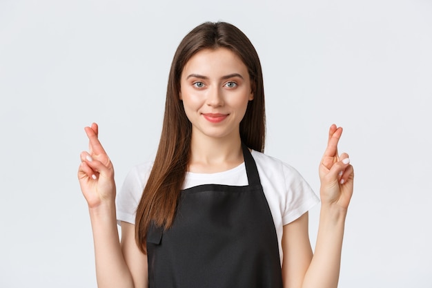Employees, job employment, small business and coffee shop concept. Confident hopeful saleswoman in black apron making wish. Barista cross fingers good luck and smiling.