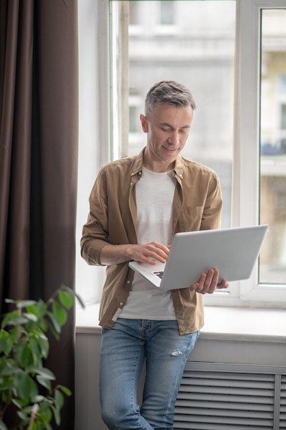 Employment. Smiling middle aged man standing with back to window in room during day holding laptop working