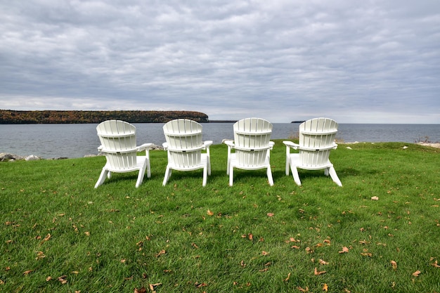 Empty adirondack chairs at grassy lakeshore against cloudy sky