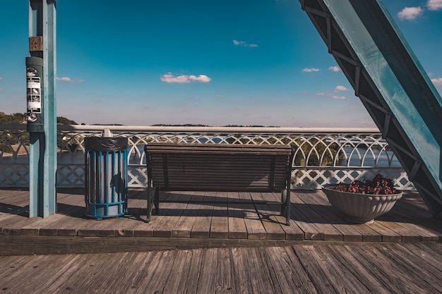 Photo empty bench by garbage bin on floorboard against sky