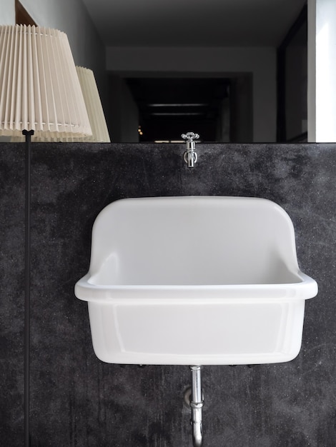 Empty big white vintage medical wall mount bathroom sink on black concrete wall decoration near big mirror and standing light lamp in the bathroom vertical style White farmhouse sink bathtub design