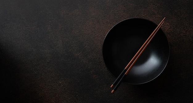 Empty black ceramic bowl with chopsticks on dark brown stone background, top view, copy space