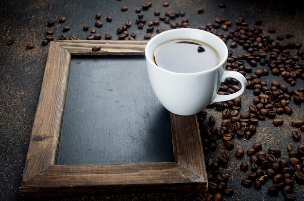 Empty black frame with White cup of black coffee and coffee beans on dark concrete background. The concept of hot drinks,  Invigorating breakfast. copy space.