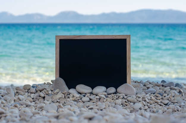 Empty blackboard on a pebble beach with turquoise water and mountains in the background